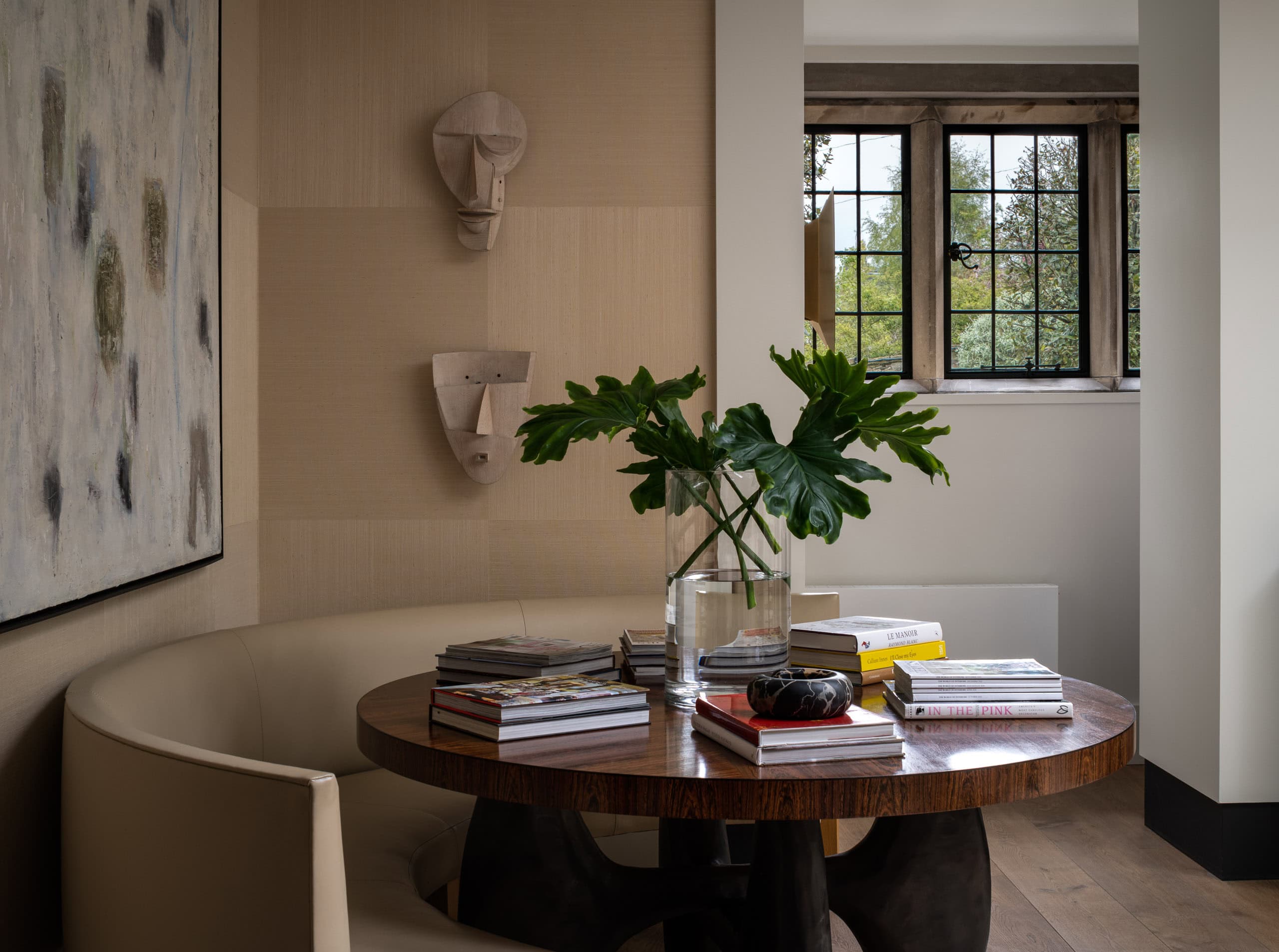 Modern interior with a round wooden table, books, and a large leafy plant near a window.