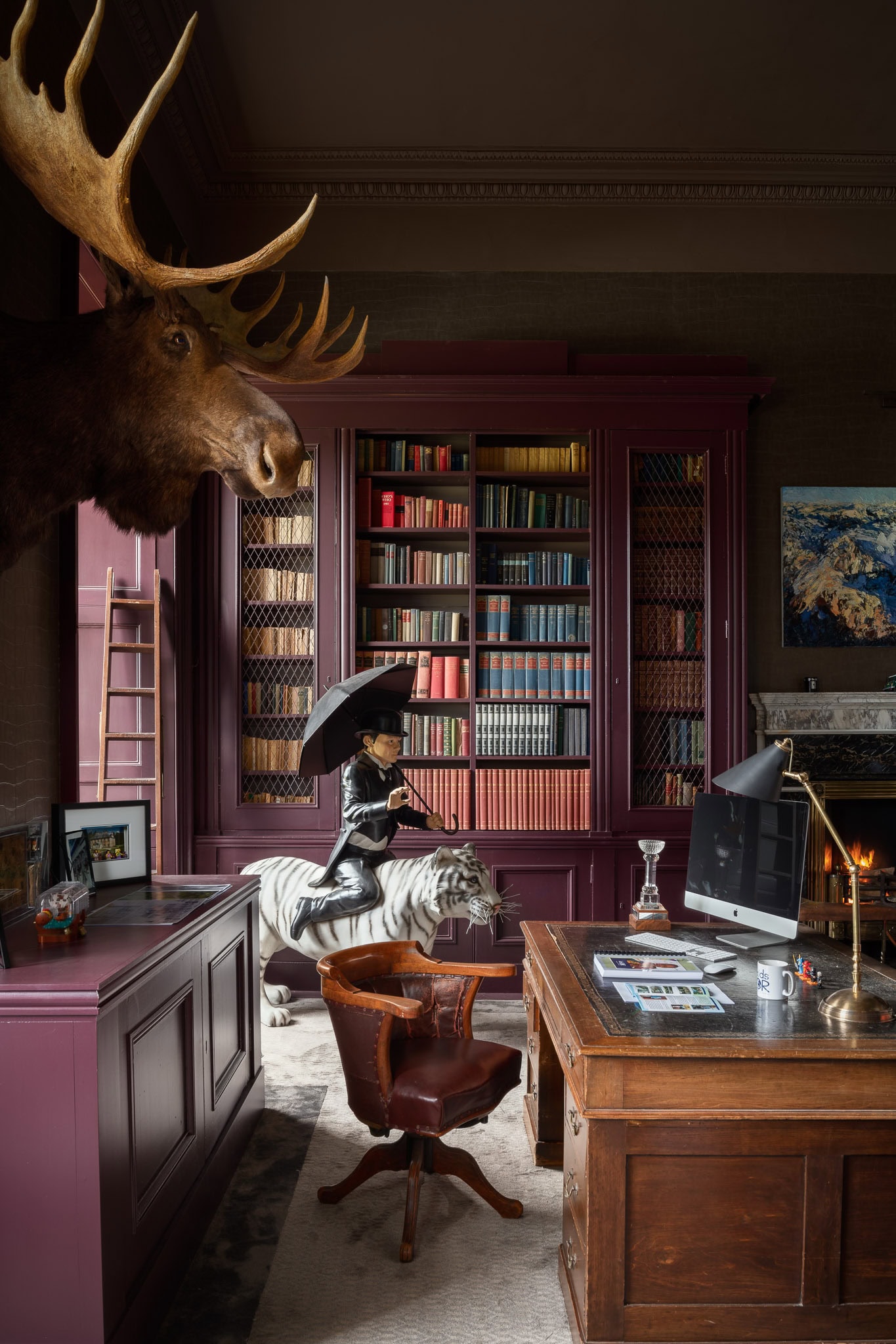 A deer head mounted on the wall in a stylish, vintage office with a large wooden desk, bookshelves,.