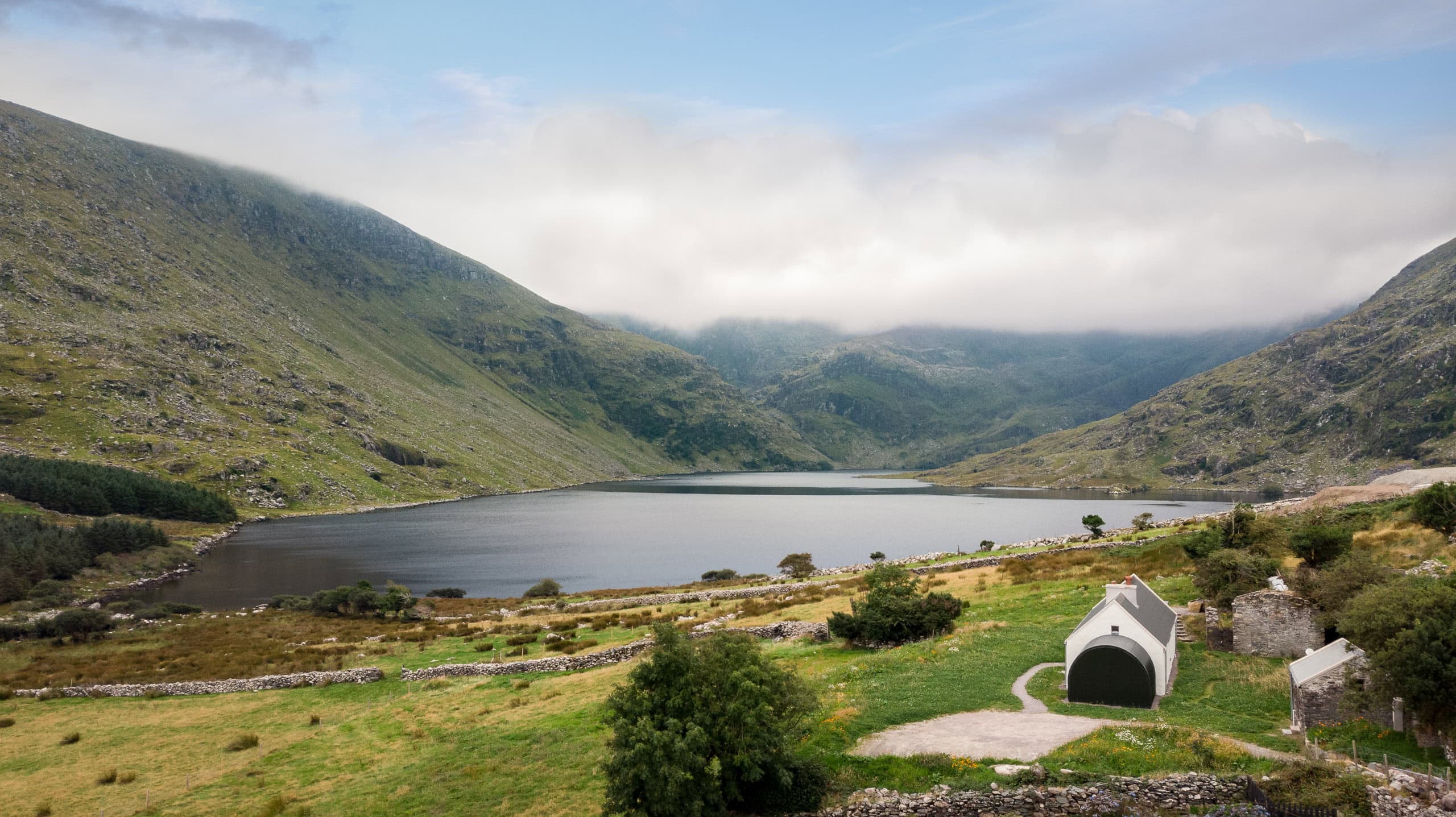 Remote lochside cottage nestled within the mountainous landscape of County Kerry featuring rolling green hills, calm water, and low cloud cover.