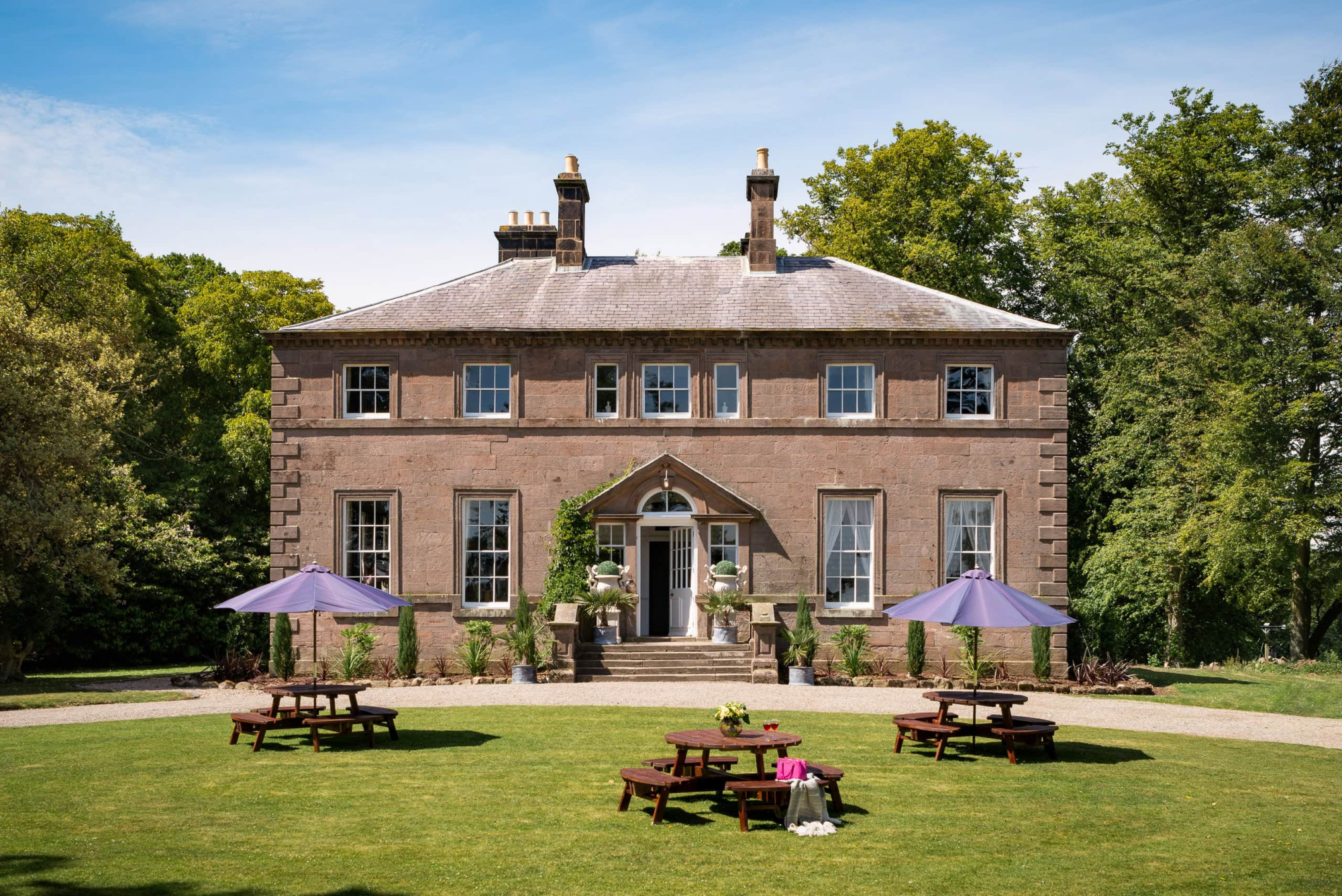 Elegant historic mansion with lush gardens and outdoor seating in a scenic countryside setting.
