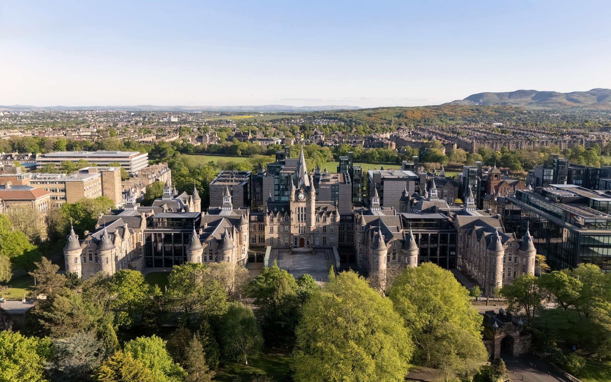 Aerial architectural view of the Edinburgh Futures Institute by Bennetts Associates, surrounded by historic Edinburgh buildings, landscaped grounds, and distant hills under clear skies.