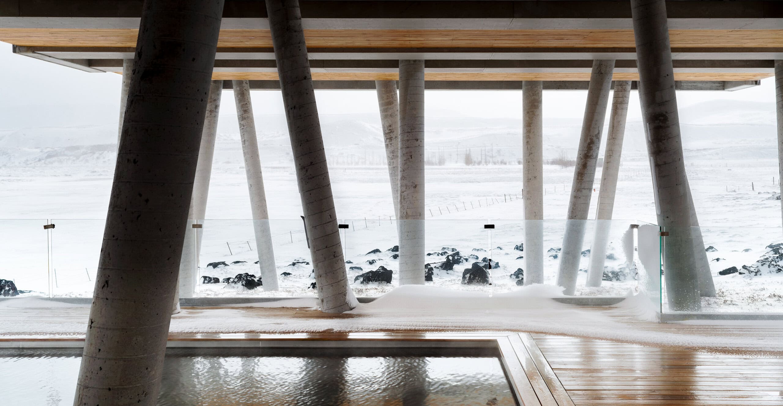 Geothermal swimming pool beneath the Ion Adventure Hotel in Iceland featuring angled concrete columns, timber decking, and a snow-covered volcanic landscape.