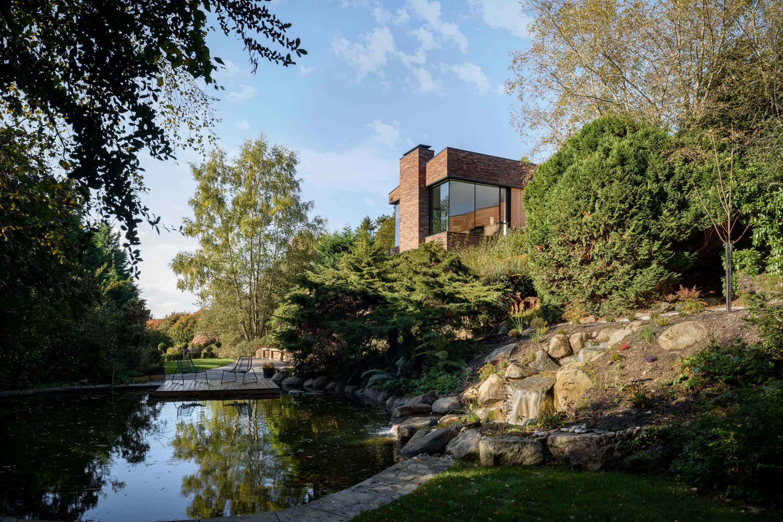 Contemporary Pond House by Technique Studio overlooking a landscaped garden pond with natural stone waterfalls, mature planting, and expansive glazing.