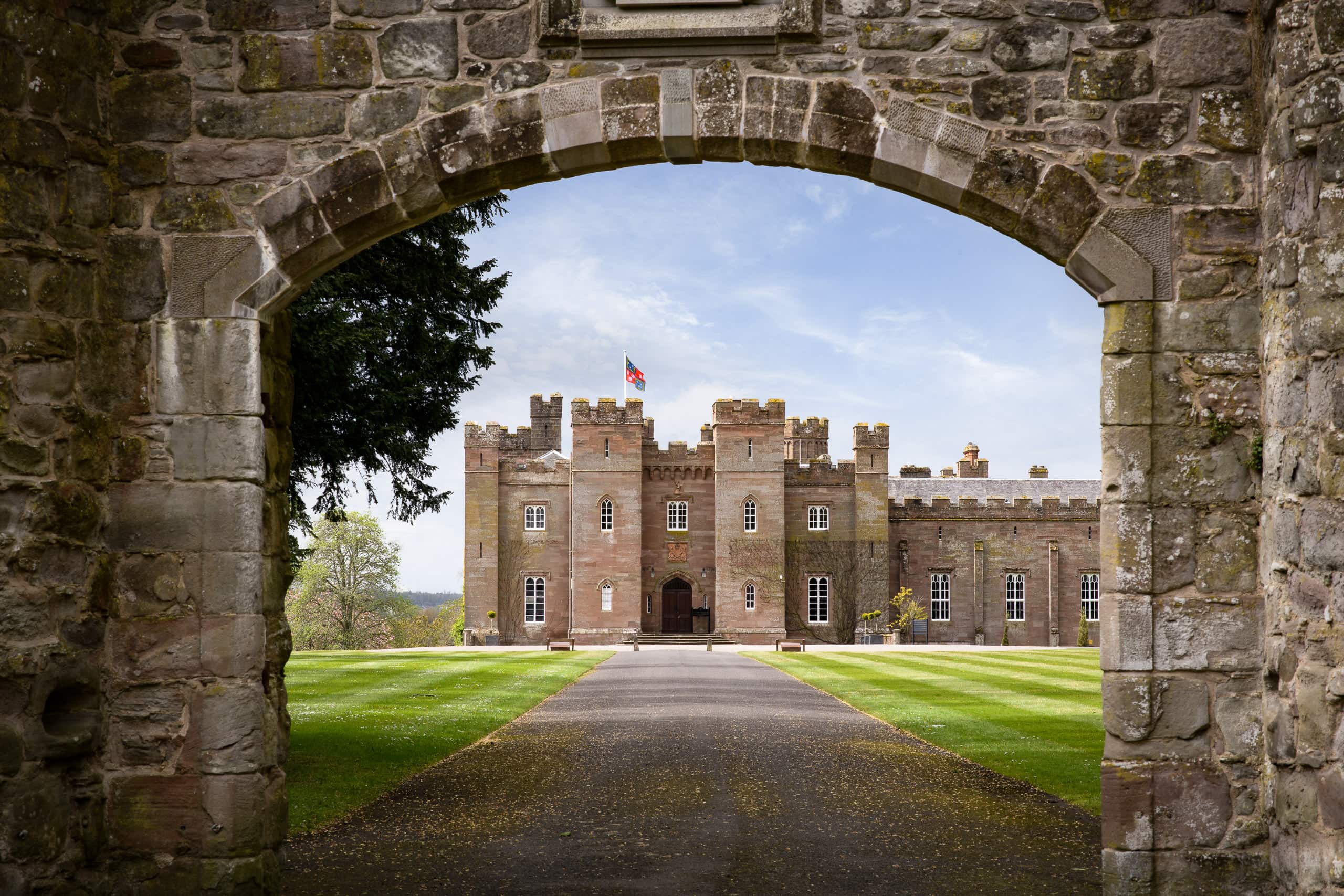 Historic castle viewed through stone archway with lush grounds and blue sky.